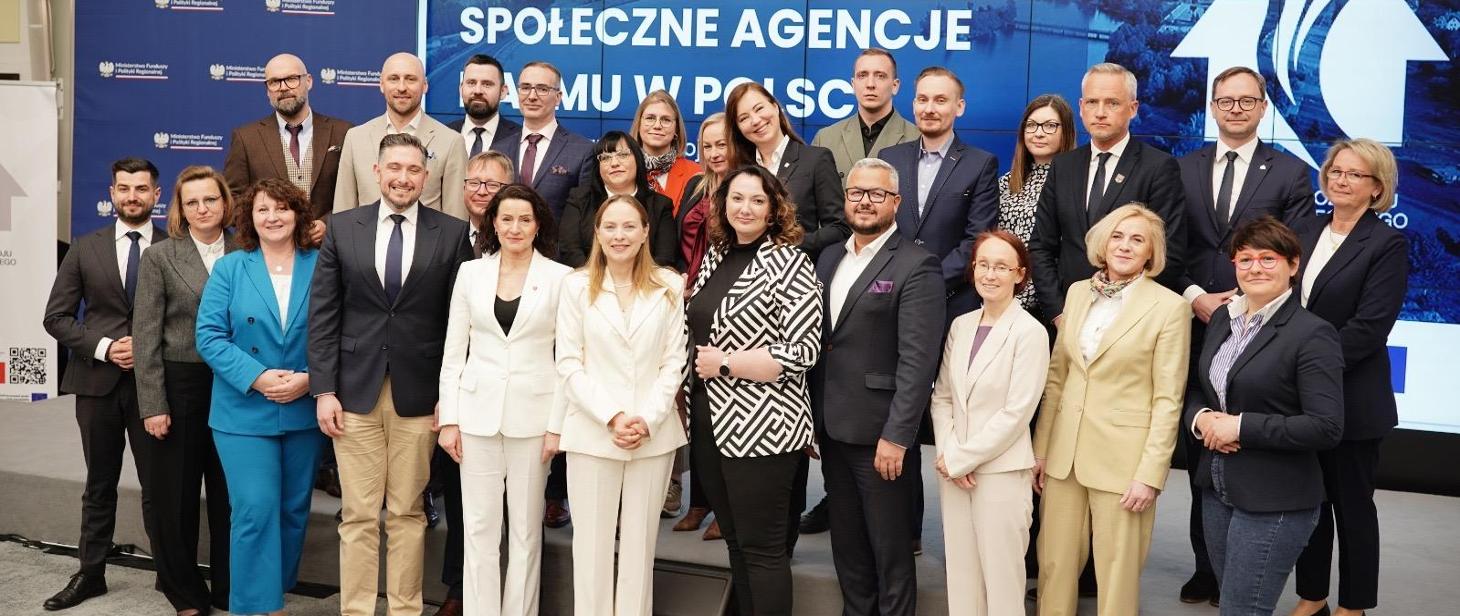 Grupa osób pozuje do wspólnego zdjęcia podczas wydarzenia dotyczącego Społecznych Agencji Najmu w Polsce. W tle widać ekran z nazwą wydarzenia i logotypami Ministerstwa Funduszy i Polityki Regionalnej